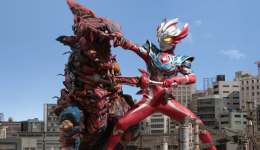 Ultraman Taiga, on the right, engages a red kaiju, on the left. They tower over the Japanese city they battle within.