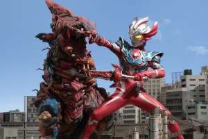 Ultraman Taiga, on the right, engages a red kaiju, on the left. They tower over the Japanese city they battle within.