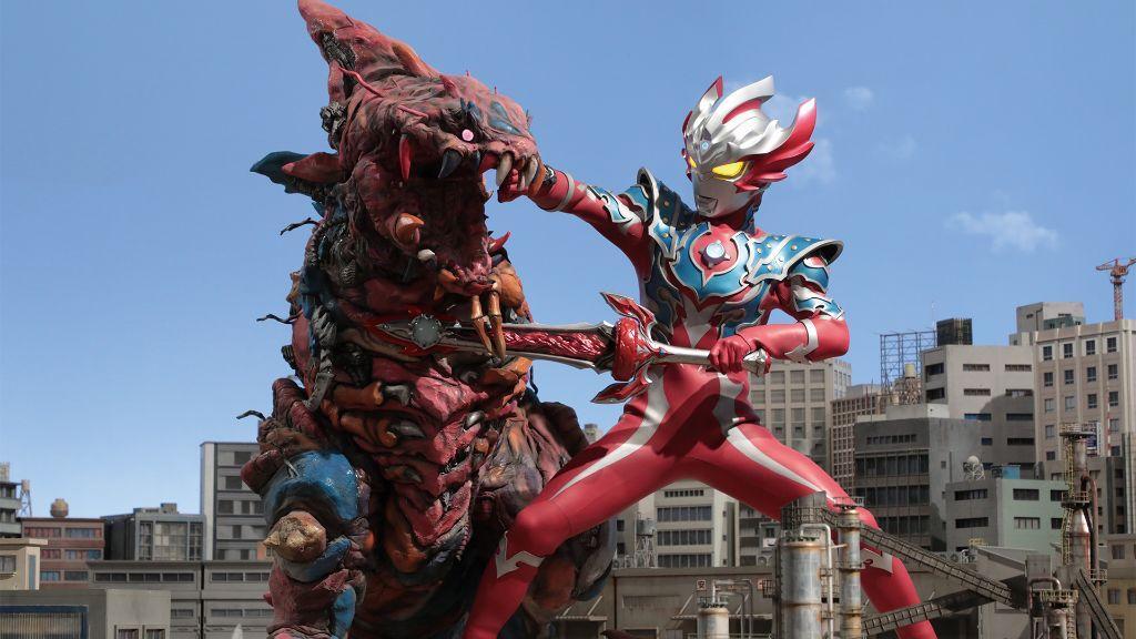 Ultraman Taiga, on the right, engages a red kaiju, on the left. They tower over the Japanese city they battle within.