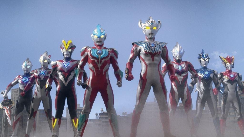 A group of Ultramen, led by Ultraman Taiga (Front Right) stand heroically against a clear blue sky.