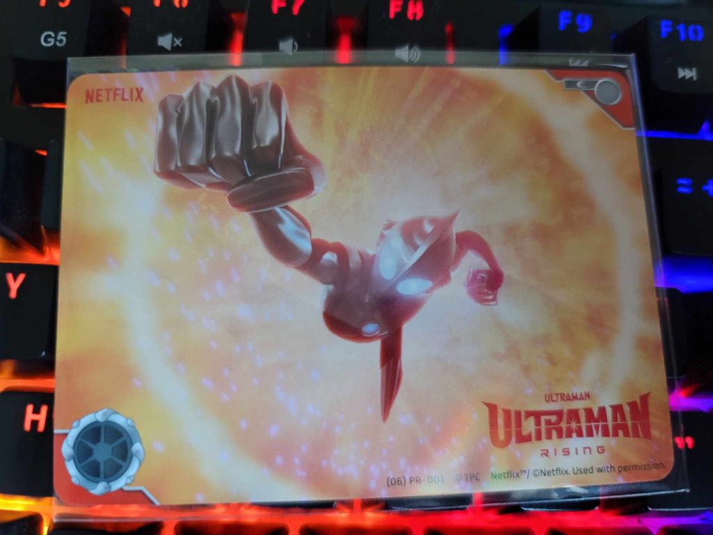 A photo of the promotional card for the ULTRAMAN CARD GAME. The card features a screenshot from ULTRAMAN RISING, with Ultraman recreating the iconic transformation pose of the original Ultraman.