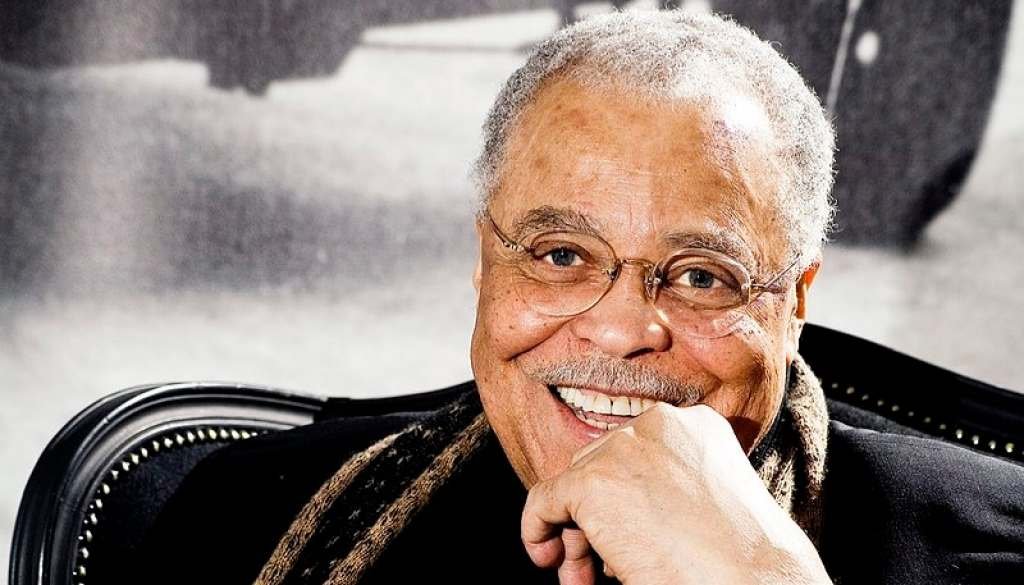 A photo of James Earl Jones sitting in a chair with his left hand by his chin, smiling for the camera, wearing a black suit and a brown scarf in 2010