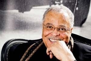 A photo of James Earl Jones sitting in a chair with his left hand by his chin, smiling for the camera, wearing a black suit and a brown scarf in 2010