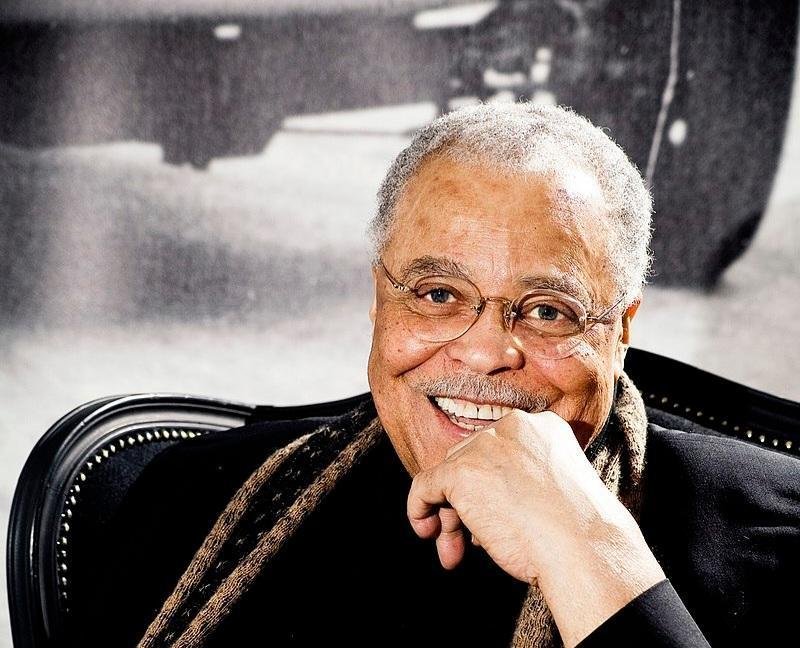 A photo of James Earl Jones sitting in a chair with his left hand by his chin, smiling for the camera, wearing a black suit and a brown scarf in 2010