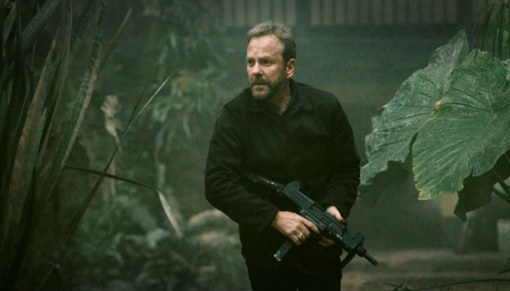 A man holding a firearm while treading between a field of tall greens.