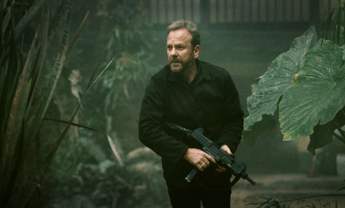 A man holding a firearm while treading between a field of tall greens.