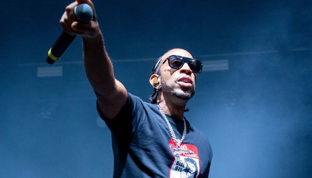 A man in sunglasses and a dark t-shirt stands before a stadium audience with his arm extended and holding a microphone.