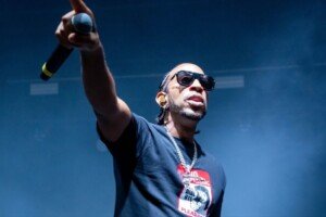 A man in sunglasses and a dark t-shirt stands before a stadium audience with his arm extended and holding a microphone.