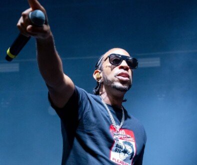 A man in sunglasses and a dark t-shirt stands before a stadium audience with his arm extended and holding a microphone.