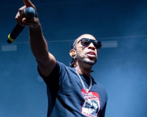 A man in sunglasses and a dark t-shirt stands before a stadium audience with his arm extended and holding a microphone.