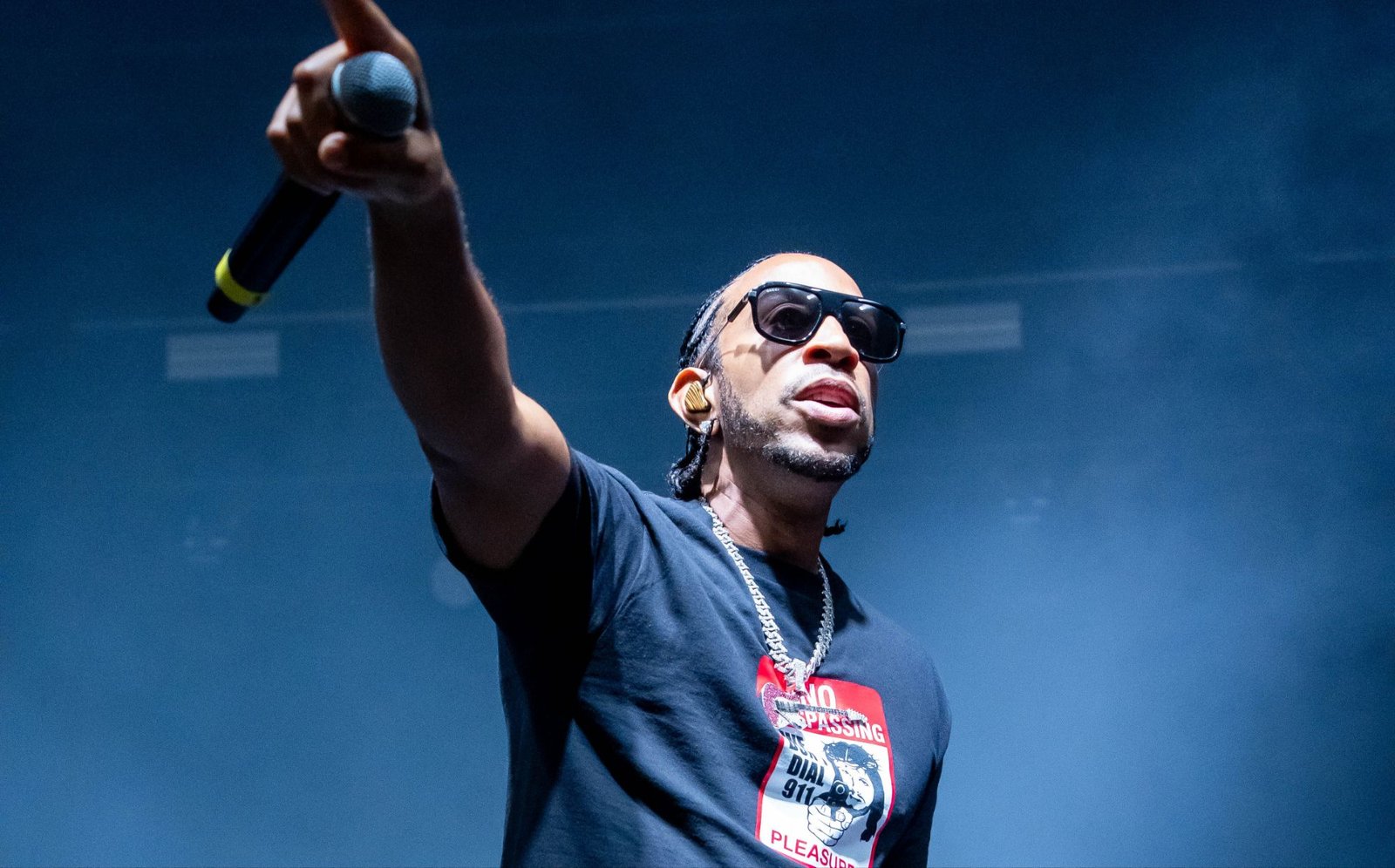 A man in sunglasses and a dark t-shirt stands before a stadium audience with his arm extended and holding a microphone.