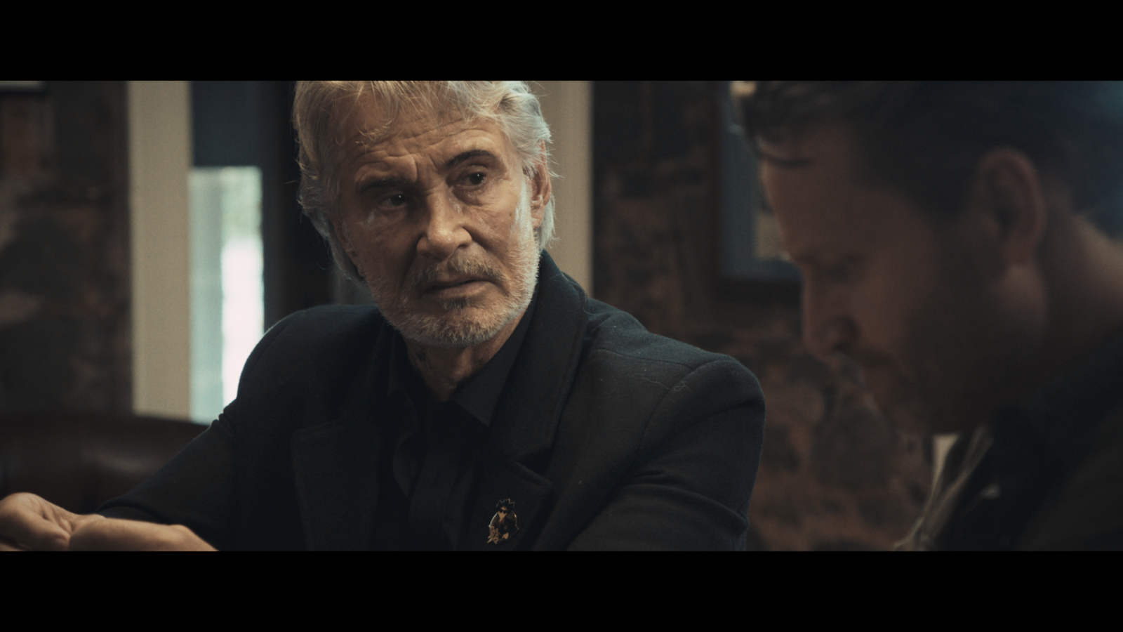 A man in a black suit turns attentively to another man next to him.